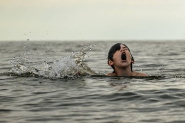 Bir çocuk kollarını kasıp gülümserken renkli bir plaj topu tutarak suda duruyor. Arkaplanda açık bir gökyüzü ve uzak kıyı şeridi yer alıyor.