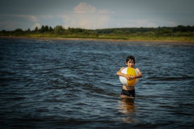 Bir çocuk kollarını kasıp gülümserken renkli bir plaj topu tutarak suda duruyor. Arkaplanda açık bir gökyüzü ve uzak kıyı şeridi yer alıyor.