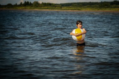 Bir çocuk kollarını kasıp gülümserken renkli bir plaj topu tutarak suda duruyor. Arkaplanda açık bir gökyüzü ve uzak kıyı şeridi yer alıyor.