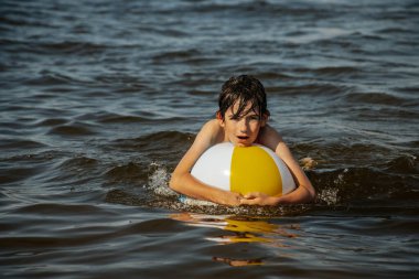 Bir çocuk kollarını kasıp gülümserken renkli bir plaj topu tutarak suda duruyor. Arkaplanda açık bir gökyüzü ve uzak kıyı şeridi yer alıyor.
