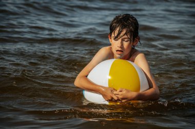 Bir çocuk kollarını kasıp gülümserken renkli bir plaj topu tutarak suda duruyor. Arkaplanda açık bir gökyüzü ve uzak kıyı şeridi yer alıyor.