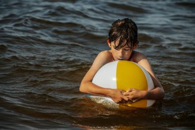 Bir çocuk kollarını kasıp gülümserken renkli bir plaj topu tutarak suda duruyor. Arkaplanda açık bir gökyüzü ve uzak kıyı şeridi yer alıyor.