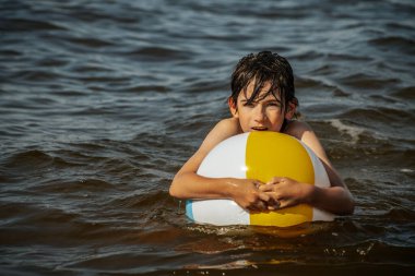 Bir çocuk kollarını kasıp gülümserken renkli bir plaj topu tutarak suda duruyor. Arkaplanda açık bir gökyüzü ve uzak kıyı şeridi yer alıyor.