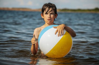 Bir çocuk kollarını kasıp gülümserken renkli bir plaj topu tutarak suda duruyor. Arkaplanda açık bir gökyüzü ve uzak kıyı şeridi yer alıyor.