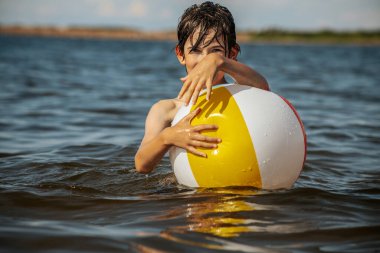 Bir çocuk kollarını kasıp gülümserken renkli bir plaj topu tutarak suda duruyor. Arkaplanda açık bir gökyüzü ve uzak kıyı şeridi yer alıyor.