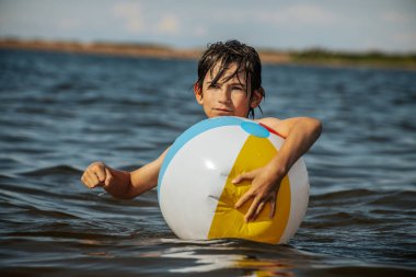 Bir çocuk kollarını kasıp gülümserken renkli bir plaj topu tutarak suda duruyor. Arkaplanda açık bir gökyüzü ve uzak kıyı şeridi yer alıyor.