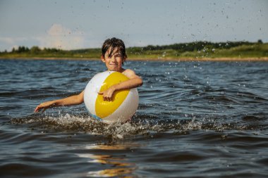 Bir çocuk kollarını kasıp gülümserken renkli bir plaj topu tutarak suda duruyor. Arkaplanda açık bir gökyüzü ve uzak kıyı şeridi yer alıyor.