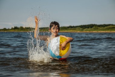 Bir çocuk kollarını kasıp gülümserken renkli bir plaj topu tutarak suda duruyor. Arkaplanda açık bir gökyüzü ve uzak kıyı şeridi yer alıyor.