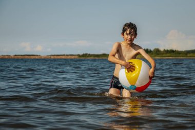 Bir çocuk kollarını kasıp gülümserken renkli bir plaj topu tutarak suda duruyor. Arkaplanda açık bir gökyüzü ve uzak kıyı şeridi yer alıyor.