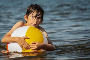 Bir çocuk kollarını kasıp gülümserken renkli bir plaj topu tutarak suda duruyor. Arkaplanda açık bir gökyüzü ve uzak kıyı şeridi yer alıyor.