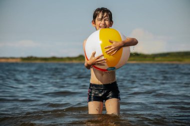 Bir çocuk kollarını kasıp gülümserken renkli bir plaj topu tutarak suda duruyor. Arkaplanda açık bir gökyüzü ve uzak kıyı şeridi yer alıyor.