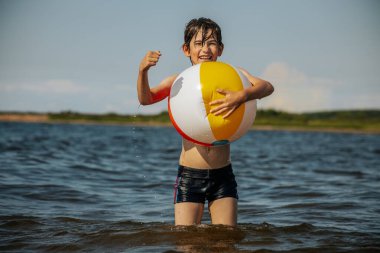 Bir çocuk kollarını kasıp gülümserken renkli bir plaj topu tutarak suda duruyor. Arkaplanda açık bir gökyüzü ve uzak kıyı şeridi yer alıyor.