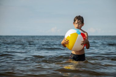 Bir çocuk kollarını kasıp gülümserken renkli bir plaj topu tutarak suda duruyor. Arkaplanda açık bir gökyüzü ve uzak kıyı şeridi yer alıyor.