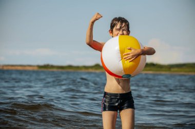 Bir çocuk kollarını kasıp gülümserken renkli bir plaj topu tutarak suda duruyor. Arkaplanda açık bir gökyüzü ve uzak kıyı şeridi yer alıyor.