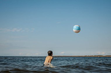 Bir çocuk kollarını kasıp gülümserken renkli bir plaj topu tutarak suda duruyor. Arkaplanda açık bir gökyüzü ve uzak kıyı şeridi yer alıyor.