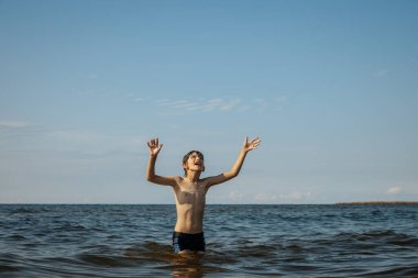 Bir çocuk kollarını kasıp gülümserken renkli bir plaj topu tutarak suda duruyor. Arkaplanda açık bir gökyüzü ve uzak kıyı şeridi yer alıyor.