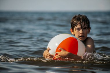 Bir çocuk suda kameraya doğru bakarken renkli bir plaj topu tutuyor. Arka plan güneşli bir gökyüzünün altında sakin bir denizi gösteriyor..