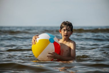 Bir çocuk suda kameraya doğru bakarken renkli bir plaj topu tutuyor. Arka plan güneşli bir gökyüzünün altında sakin bir denizi gösteriyor..