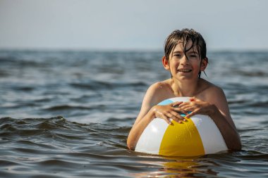 Bir çocuk suda kameraya doğru bakarken renkli bir plaj topu tutuyor. Arka plan güneşli bir gökyüzünün altında sakin bir denizi gösteriyor..
