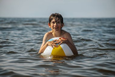 Bir çocuk suda kameraya doğru bakarken renkli bir plaj topu tutuyor. Arka plan güneşli bir gökyüzünün altında sakin bir denizi gösteriyor..