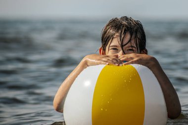 Bir çocuk suda kameraya doğru bakarken renkli bir plaj topu tutuyor. Arka plan güneşli bir gökyüzünün altında sakin bir denizi gösteriyor..