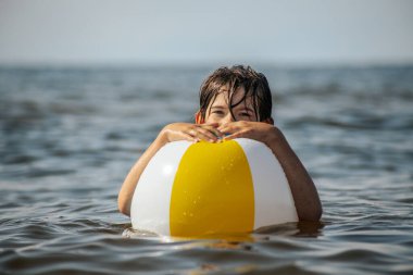 Bir çocuk suda kameraya doğru bakarken renkli bir plaj topu tutuyor. Arka plan güneşli bir gökyüzünün altında sakin bir denizi gösteriyor..