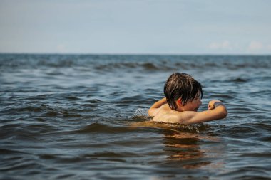 Güneşli bir günde okyanusta oynamanın heyecanını yakalayan, kolları havada suya neşeyle sıçrayan bir çocuk. Sahne, çocukluk ve yaz eğlencesinin tasasız ruhunu simgeliyor.