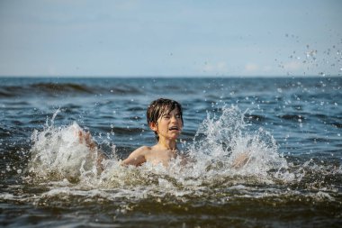 Güneşli bir günde okyanusta oynamanın heyecanını yakalayan, kolları havada suya neşeyle sıçrayan bir çocuk. Sahne, çocukluk ve yaz eğlencesinin tasasız ruhunu simgeliyor.
