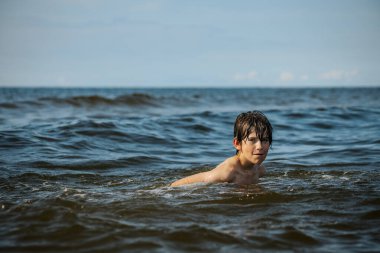 Güneşli bir günde okyanusta oynamanın heyecanını yakalayan, kolları havada suya neşeyle sıçrayan bir çocuk. Sahne, çocukluk ve yaz eğlencesinin tasasız ruhunu simgeliyor.