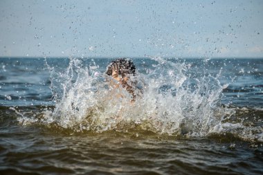 Güneşli bir günde okyanusta oynamanın heyecanını yakalayan, kolları havada suya neşeyle sıçrayan bir çocuk. Sahne, çocukluk ve yaz eğlencesinin tasasız ruhunu simgeliyor.