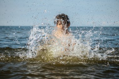 Güneşli bir günde okyanusta oynamanın heyecanını yakalayan, kolları havada suya neşeyle sıçrayan bir çocuk. Sahne, çocukluk ve yaz eğlencesinin tasasız ruhunu simgeliyor.