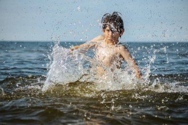 Güneşli bir günde okyanusta oynamanın heyecanını yakalayan, kolları havada suya neşeyle sıçrayan bir çocuk. Sahne, çocukluk ve yaz eğlencesinin tasasız ruhunu simgeliyor.