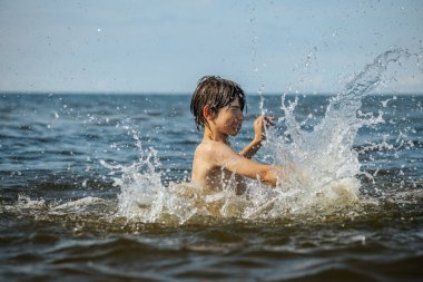 Güneşli bir günde okyanusta oynamanın heyecanını yakalayan, kolları havada suya neşeyle sıçrayan bir çocuk. Sahne, çocukluk ve yaz eğlencesinin tasasız ruhunu simgeliyor.