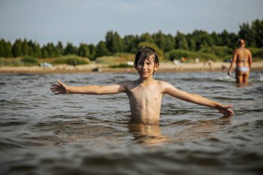 Kollarını açmış gülümseyen bir çocuk sahilde suyun içinde duruyor. Sahile gidenler arka planda berrak bir gökyüzünün altında görülebilir.