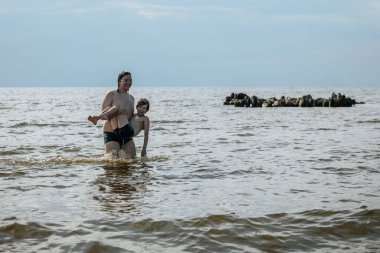 Bir ebeveyn çocuğunu okyanusta kucağına alır, ikisi de mayo giyer. Gökyüzü bulutlu ve su ufka kadar uzanıyor.
