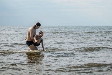 Bir ebeveyn çocuğunu okyanusta kucağına alır, ikisi de mayo giyer. Gökyüzü bulutlu ve su ufka kadar uzanıyor.