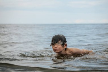 Gözlük takan bir çocuk neşeli bir ifadeyle suyun içinde duruyor, saçından damlayan su damlacıklarıyla. Sahne, bir yaz günü yüzmenin eğlencesini ve heyecanını yakalar..