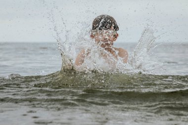 Gözlük takan bir çocuk sevinçle suya su sıçratır, heyecanla kollarını kaldırır. Sahne, çocukluk ve yaz eğlencesinin kaygısız neşesini yakalar.