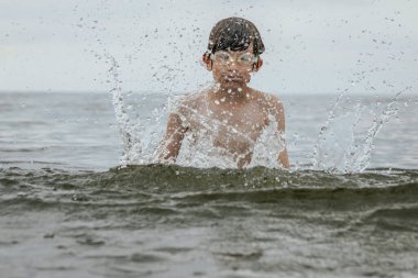 Gözlük takan bir çocuk sevinçle suya su sıçratır, heyecanla kollarını kaldırır. Sahne, çocukluk ve yaz eğlencesinin kaygısız neşesini yakalar.