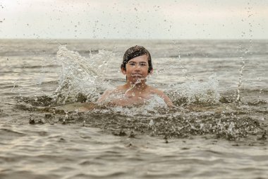 Suyun içinde çırpınan ve bağıran genç bir çocuk çevresindeki sıçramalarla ve arka planda geniş bir su birikintisiyle acı çekiyor gibi görünüyor. Sahne, aciliyet ve endişe hissi uyandırıyor.