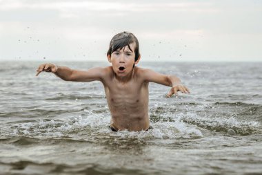 Suyun içinde çırpınan ve bağıran genç bir çocuk çevresindeki sıçramalarla ve arka planda geniş bir su birikintisiyle acı çekiyor gibi görünüyor. Sahne, aciliyet ve endişe hissi uyandırıyor.