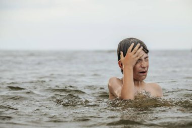 Suyun içinde çırpınan ve bağıran genç bir çocuk çevresindeki sıçramalarla ve arka planda geniş bir su birikintisiyle acı çekiyor gibi görünüyor. Sahne, aciliyet ve endişe hissi uyandırıyor.