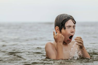 Suyun içinde çırpınan ve bağıran genç bir çocuk çevresindeki sıçramalarla ve arka planda geniş bir su birikintisiyle acı çekiyor gibi görünüyor. Sahne, aciliyet ve endişe hissi uyandırıyor.