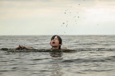 Suyun içinde çırpınan ve bağıran genç bir çocuk çevresindeki sıçramalarla ve arka planda geniş bir su birikintisiyle acı çekiyor gibi görünüyor. Sahne, aciliyet ve endişe hissi uyandırıyor.