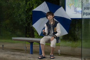 Bir çocuk mavi ve beyaz bir şemsiyeyle oturur, dalgın dalgın bakar. Karanlık arka plan ve düşünceli ifadesi sessiz bir yansıma yaratıyor..