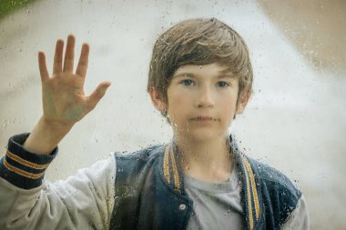 Elleri sırılsıklam olmuş pencereye uzanmış neşeli bir çocuk, ışıl ışıl gülümsüyor. Camdaki damlacıklar ve bulanık arka plan neşeli ve canlandırıcı bir atmosfer yaratıyor..