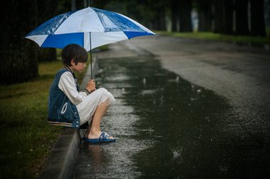 Yağmurlu bir günde, bir su birikintisinde suya dokunan, mavi beyaz bir şemsiyeyle kaldırımda oturan bir çocuk. Meraklı ifadesi ve yağmurlu ortam bir merak ve keşif hissi yaratıyor..