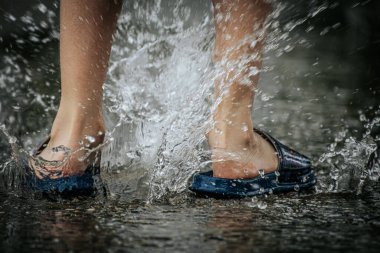 Su birikintilerinin arasından süzülen ayaklar dinamik bir su patlaması yaratır. Mavi sandalet giyen kişi, neşeli ve kaygısız anı vurguluyor..