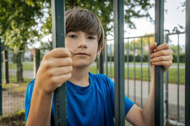 Mavi gömlekli genç bir çocuk parkta dikilirken metal parmaklıkları kavrıyor ve doğrudan kameraya bakıyor. Arkaplanda yeşil ağaçlar ve bir çit var.