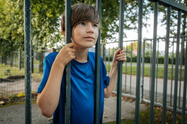 Mavi gömlekli genç bir çocuk parkta dikilirken metal parmaklıkları kavrıyor ve doğrudan kameraya bakıyor. Arkaplanda yeşil ağaçlar ve bir çit var.